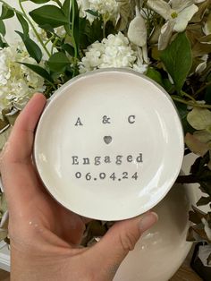 a person holding a white plate with the words and initials engraved on it in front of flowers