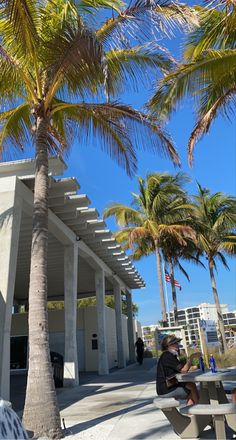 two people sitting at a picnic table under palm trees in front of a white building