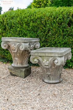 two cement planters sitting on top of a gravel ground next to green bushes and shrubbery