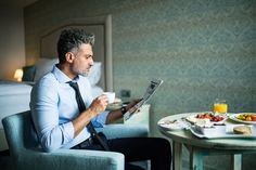 a man sitting at a table reading a newspaper and eating breakfast in a hotel room