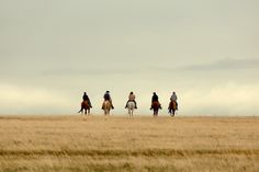 four people are riding horses in an open field with tall grass and cloudy sky behind them