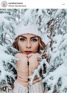 a woman wearing a hat and sweater standing in front of snow covered trees with her hands on her face