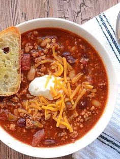 a white bowl filled with chili and cheese next to a piece of bread on top of a wooden table