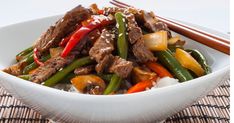 a white bowl filled with stir fry beef and vegetables next to chopsticks on a mat