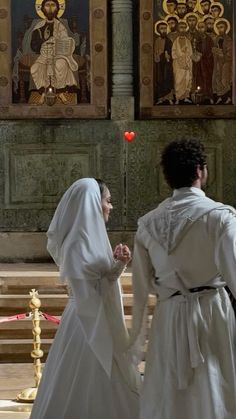 two people dressed in white standing next to each other at the alter with paintings on the wall behind them