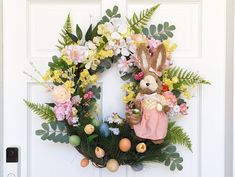 a wreath with flowers, eggs and an easter bunny on it is hanging from the front door