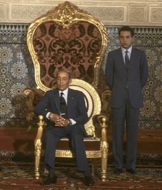 two men in suits and ties are sitting on a gold chair with an ornately decorated wall behind them