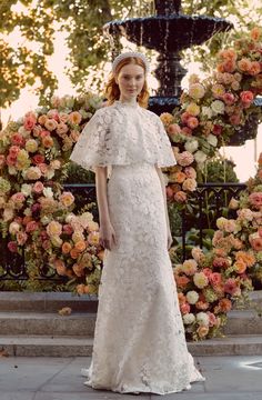 a woman standing in front of flowers wearing a wedding dress with ruffles on the shoulders