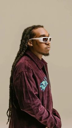 a man with dreadlocks wearing sunglasses and a corded jacket standing in front of a gray background