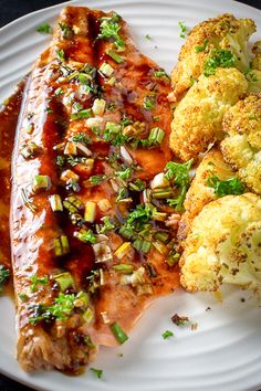 a white plate topped with meat covered in sauce and veggies next to cauliflower