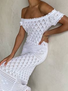 a woman leaning against a wall wearing a white crochet dress and gold jewelry