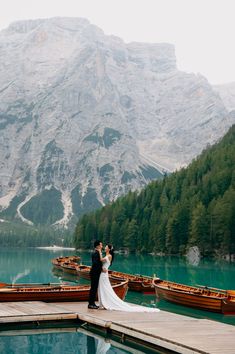 a bride and groom standing on a dock in front of some boats with mountains in the background