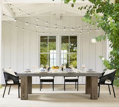 an outdoor dining table with white chairs and lights strung from the ceiling over it, surrounded by greenery