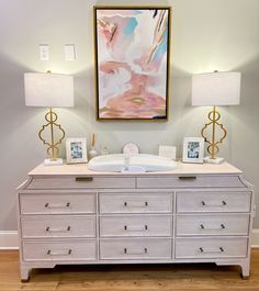 a white dresser with two lamps on top and a painting hanging above it in the corner