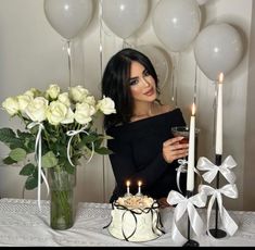 a woman sitting at a table with a cake and candles in front of her, surrounded by balloons