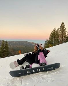 two snowboarders sitting in the snow on top of a hill at sunset with trees and mountains in the background