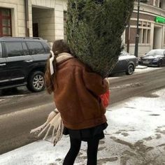 a woman walking down the street carrying a christmas tree on her head and wearing a brown jacket