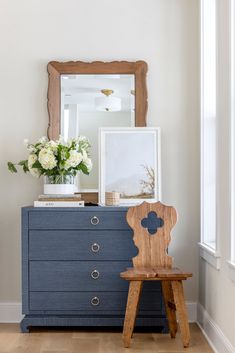 a wooden chair sitting next to a blue dresser with flowers on top and a mirror above it