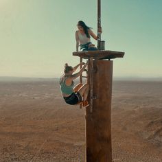 two women climbing up the side of a tall wooden structure in the middle of nowhere