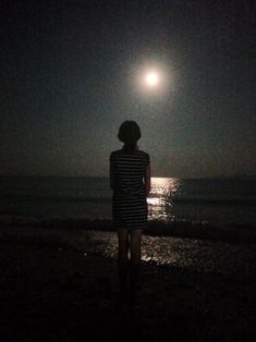 a person standing on the beach at night looking out into the ocean with the moon in the background