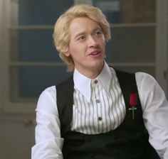 a man with blonde hair wearing a black vest and white shirt is holding a red rose in his right hand