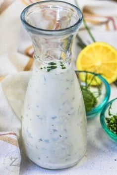 a glass pitcher filled with dressing next to bowls of herbs and lemons on a table