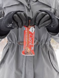 a person wearing gloves holding a bar of candy in their hands and snow on the ground behind them