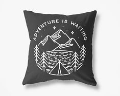 a black pillow with the words adventure is waiting in front of it and trees around it