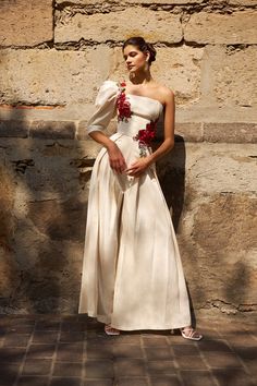 a woman standing in front of a stone wall wearing a white dress with flowers on it