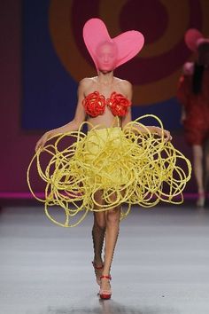 a woman walking down a runway wearing a pink hat and yellow dress with red flowers on it