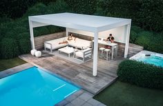 two people sitting at a table on a patio next to a swimming pool and covered with white furniture