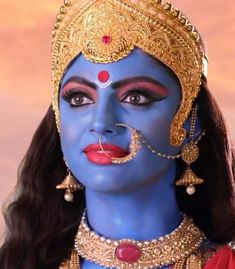 a close up of a person wearing blue makeup and gold jewelry on their face with an orange sky in the background