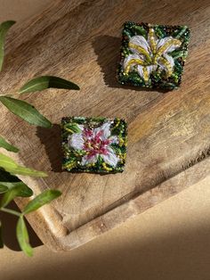 two square beads with flowers on them sitting on a wooden surface next to a plant