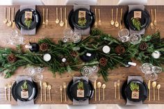 the table is set with black plates and silverware, pine cones, greenery, and candles