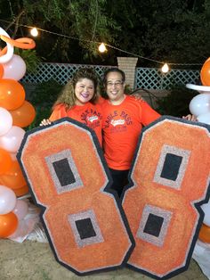 two people standing in front of an orange and black number sign with balloons around them