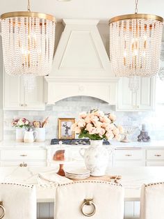 a white kitchen with chandeliers hanging from the ceiling and flowers on the counter