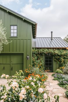 a green house with white flowers in the foreground and an olive green building behind it