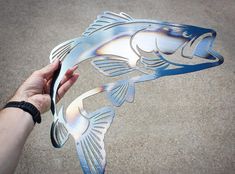 a hand holding a metal fish cutout on the ground with it's mouth open