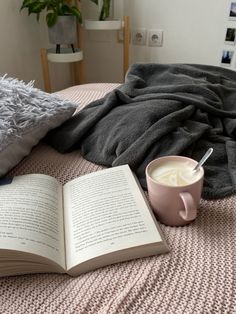 an open book sitting on top of a bed next to a cup of coffee and blanket