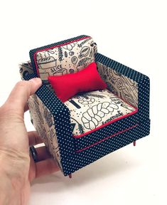 a hand is holding a miniature chair made out of fabric with red pillows on it