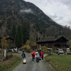 several people walking down a dirt road in front of a mountain side house with cars parked on the other side