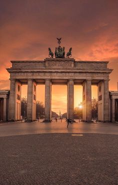 the sun is setting at the end of an arch with statues on top and people walking around