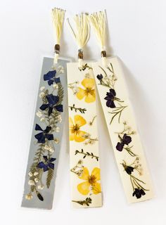 three bookmarks with flowers on them are hanging from the wall and decorated with tassels