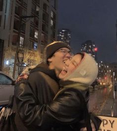 a man and woman kissing on the street in front of tall buildings at night time