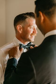 a man adjusting his bow tie while looking into another man's eyes and smiling