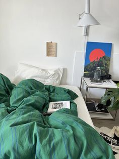 an unmade bed with green comforter in a white room next to a painting on the wall