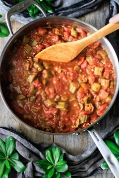 a wooden spoon in a large pot filled with vegetable stew and garnished with basil leaves