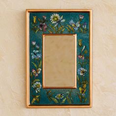 a mirror mounted to the side of a wall with flowers painted on it and gold trim