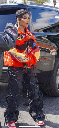 a woman in an orange jacket and black pants is standing next to a car with her hand on her hip