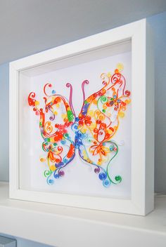 a colorful butterfly in a white frame sitting on top of a shelf next to a blue wall